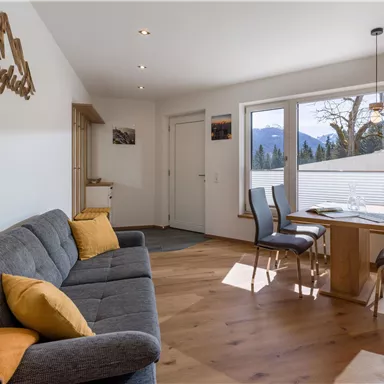 Ein modernes Wohnzimmer mit einer grau-gestreiften Couch und einem Holztisch. Große Fenster bieten einen Blick auf die Berge und bringen viel Tageslicht herein.