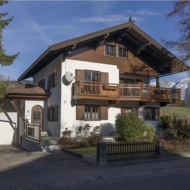A beautiful house in traditional Alpine style with balconies and a well-maintained garden. In the background, there are trees and a blue sky.