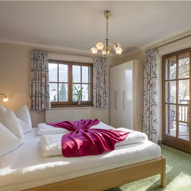 A cozy bedroom with a large bed and two pillows. The windows let in plenty of light and lead to a balcony.