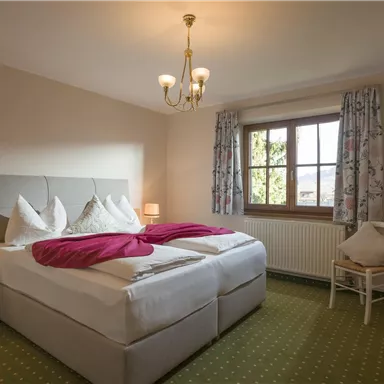 A cozy bedroom with two beds and white bedcovers. The room has large windows, a carpeted floor, and pleasant lighting.