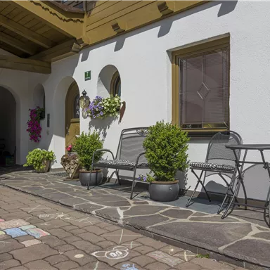 An inviting outdoor area with seating and blooming plants. The bright facade and the paved path create a cozy atmosphere.