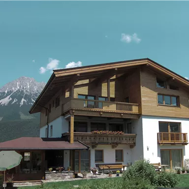 Ein modernes Haus mit Holzelementen und Balkonen in bergiger Umgebung. Im Hintergrund sind schneebedeckte Berge und ein klarer Himmel zu sehen.