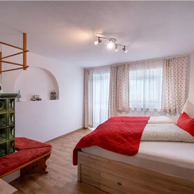 A cozy bedroom with a large bed and red pillows. The room has a wooden floor and bright windows with curtains.