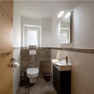 A modern bathroom with a white sink and a toilet. There are coverings in light and dark shades as well as a window for natural light.