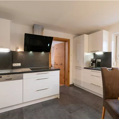 A modern kitchen with white furniture and a dark countertop. There is a dining table and large windows that let in plenty of light.