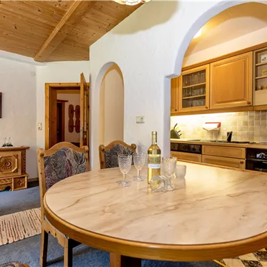 An inviting dining area with a round wooden table and cozy chairs. In the background, there is a well-equipped kitchen and bright walls.