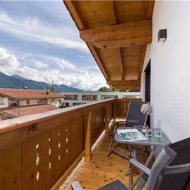A cozy balcony with a wooden railing and two chairs. In the background, mountains and a picturesque landscape are visible.
