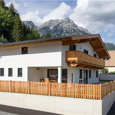 A modern house with a wooden balcony and a beautiful garden. In the background, impressive mountains and blue sky can be seen.