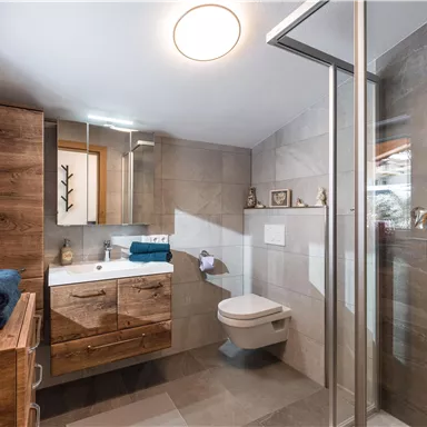 A modern bathroom with wooden details and modern tiles. It features a shower, a toilet, and a sink.