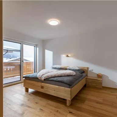 A bright bedroom with a wooden bed and two pillows. Large windows offer a view of a terrace.