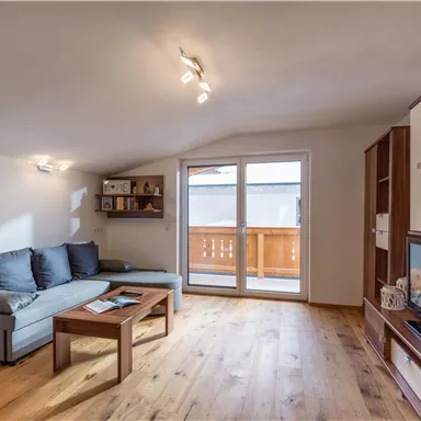 A cozy living room with a gray sofa and a wooden table. Large windows provide plenty of natural light and access to the balcony.