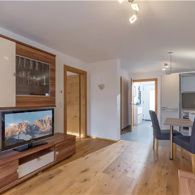 A cozy living room with modern furniture design and a television. The room features bright colors and an open access to the kitchen.