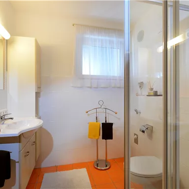 Ein helles Badezimmer mit einer Dusche und einem Waschbecken. Handtücher in Gelb und Schwarz hängen am Handtuchhalter.