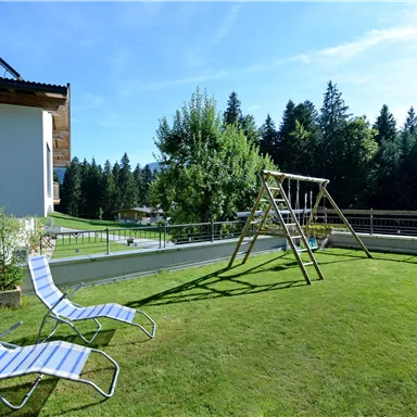 Ein schöner Garten mit einem Spielplatz und modernen Liegen. Im Hintergrund sind Bäume und ein klarer Himmel zu sehen.