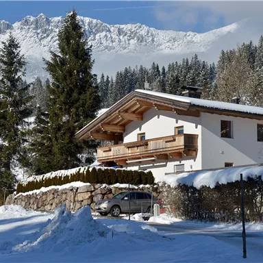 A cozy house in the mountains, surrounded by snow-covered fir trees. The majestic mountains are visible in the background.