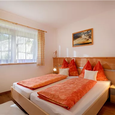 Ein gemütliches Schlafzimmer mit zwei Betten, orangefarbener Bettwäsche und Kissen. Große Fenster sorgen für viel Tageslicht und eine angenehme Atmosphäre.