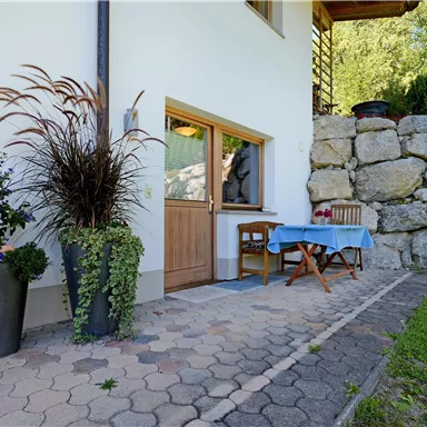 Ein einladender Eingangsbereich mit einer kleinen Sitzecke und Blumengefäßen. Steinmauer im Hintergrund und grüne Rasenfläche schaffen eine gemütliche Atmosphäre.