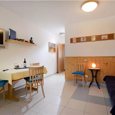 Ein helles Wohnzimmer mit einem Tisch, Stühlen und einem Sofa. An der Wand hängen Bilder und ein Fernseher ist montiert.