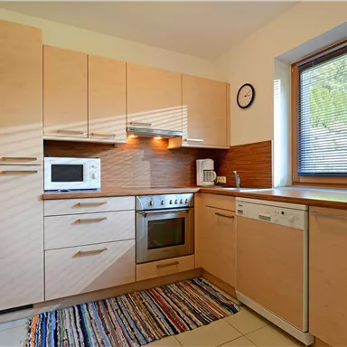 Eine moderne Küche mit hellen Holzschränken und einer Kochzone. Ein Fenster lässt viel Tageslicht herein und ein Teppich sorgt für Gemütlichkeit.