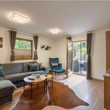 A cozy living room with a sofa and an armchair. Large windows provide a view outside and plenty of natural light.