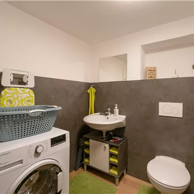 A modern bathroom with a washing machine, a sink, and a bright wall design. A laundry basket and personal care products are also visible.