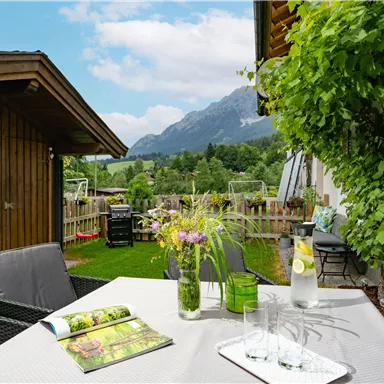 A cozy outdoor area with a table, colorful flowers, and a view of the mountains. In the background, there is a garden and a wooden shed.