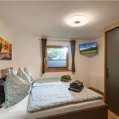 A cozy bedroom with a double bed, pillows, and clean towels. A window offers a view outside, and a television is mounted on the wall.