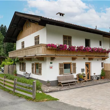 A charming house with a balcony and colorful flowers. The surroundings are green and the street passes by.