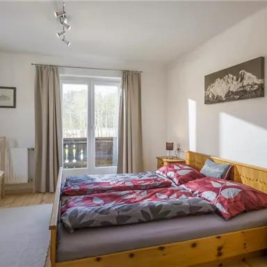 Ein helles Schlafzimmer mit einem Doppelbett und rot-grauer Bettwäsche. Es gibt Holzkommoden und große Fenster, die Tageslicht hereinlassen.