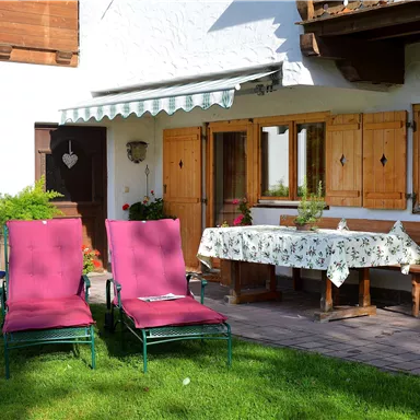 Eine gemütliche Terrasse mit zwei roten Liegestühlen und einem blühenden Tisch. Im Hintergrund sind Holzfenster und grüne Pflanzen zu sehen.