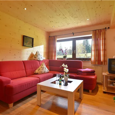 Ein gemütliches Wohnzimmer mit einem roten Sofa und einem weißen Tisch. Große Fenster sorgen für viel Tageslicht und einen Blick ins Grüne.