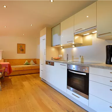 Eine moderne Küche mit hellen Schränken und einer Arbeitsfläche aus Stein. Im Hintergrund befindet sich ein gemütlicher Essbereich mit einem Tisch und Stühlen.