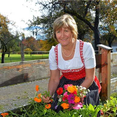Eine Frau in traditioneller Tracht pflückt Blumen in einem Garten. Im Hintergrund sind Bäume und eine Wiese zu sehen.