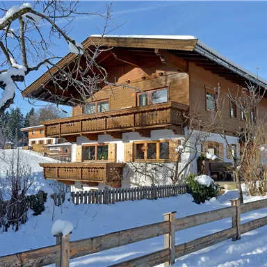 Ein gemütliches Holzhaus im Schnee, umgeben von Bäumen. Die Balkone sind schön dekoriert, und die Umgebung ist winterlich und ruhig.