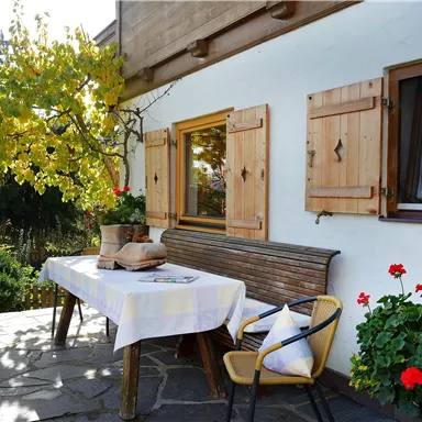 Eine einladende Terrasse mit einem Tisch und Stühlen im Grünen. Die Fensterläden sind aus Holz und es blühen rote Blumen.