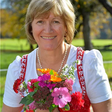 Eine lächelnde Frau trägt ein traditionelles Kleid und hält einen bunten Blumenstrauß. Im Hintergrund sind eine grüne Wiesenlandschaft und Bäume zu sehen.