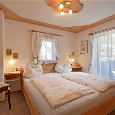 Ein gemütliches Schlafzimmer mit zwei Betten, hellen Vorhängen und warmen Holzdetails. Neben dem Bett steht ein kleiner Tisch und ein Stuhl.