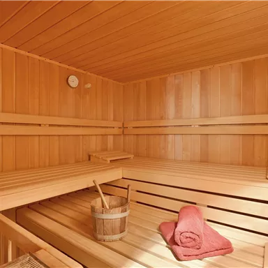 Eine gemütliche Sauna aus hellem Holz mit Bänken und einem Eimer. Ein rotes Handtuch liegt auf der Bank.
