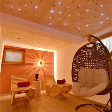 Ein gemütlicher Raum mit einem hängenden Schaukelstuhl und zwei hölzernen Sesseln. Die Wände sind in warmen Farben gehalten und eine stimmungsvolle Beleuchtung glitzert an der Decke.