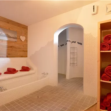 Ein heller Wellnessbereich mit einer Badewanne und roten Handtüchern. Im Hintergrund sind Holzverkleidungen und ein Durchgang zu weiteren Räumen sichtbar.