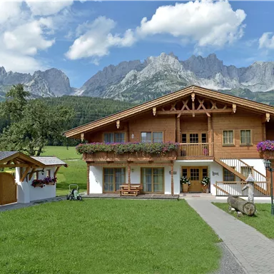 Ein wunderschönes Holzhaus inmitten einer grünen Wiese mit bunten Blumen. Im Hintergrund erheben sich majestätische Berge unter einem blauen Himmel.