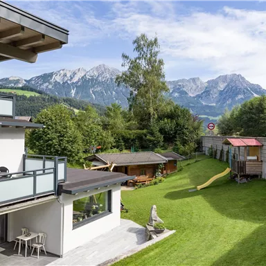 Ein schöner Garten mit einer Rasenfläche und Spielmöglichkeiten für Kinder. Im Hintergrund sind beeindruckende Berge und eine klare blaue Himmel zu sehen.