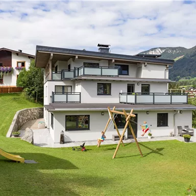 Ein modernes Haus mit mehreren Balkonen und einem großen Garten. Im Garten gibt es eine Schaukel und eine Rutsche, umgeben von Bergen und einem blauen Himmel.