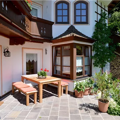 A beautiful outdoor area with a wooden table and benches. Surrounded by green plants and a bright building with windows.