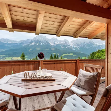 A cozy wooden balcony with a round table and chairs. In the background, the majestic mountains and a clear landscape can be seen.