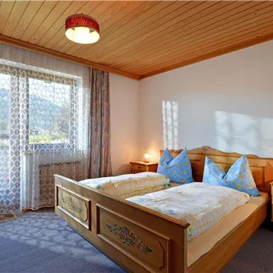 Ein helles Schlafzimmer mit einem Doppelbett und zwei blauen Kissen. Große Fenster bieten einen Blick ins Freie und lassen viel Licht herein.