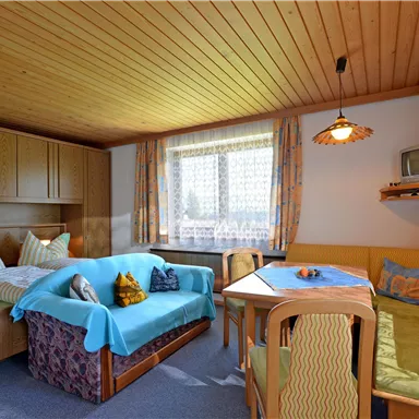 Ein gemütlicher Wohnraum mit einem Doppelbett, einer Couch und einem Essbereich. Große Fenster sorgen für viel Tageslicht.