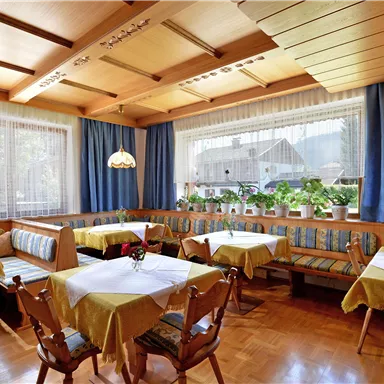 Ein gemütliches Restaurant mit Holzmöbeln und freundlich gedeckten Tischen. Durch die Fenster ist viel Licht und eine grüne Aussicht sichtbar.