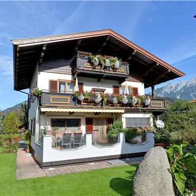 Ein charmantes, zweistöckiges Haus mit Blumenbalkonen und einem schönen Garten. Im Hintergrund sind Berge und ein klarer Himmel zu sehen.