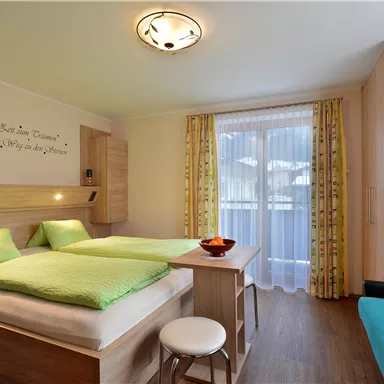 A cozy room with two beds and green cushions. The room features a balcony and a small table.